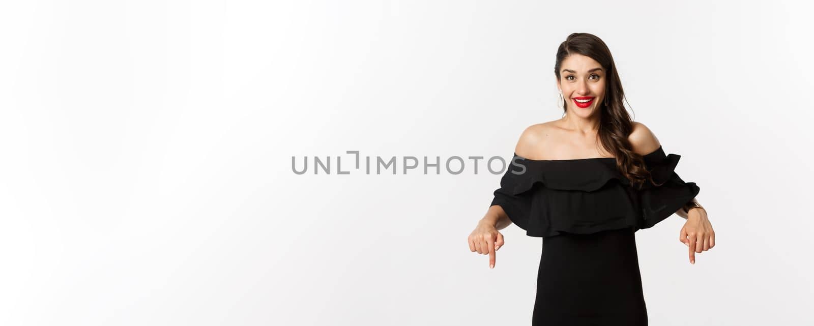 Fashion and beauty. Elegant woman in black dress pointing fingers down, showing promo and smiling, standing over white background by Benzoix