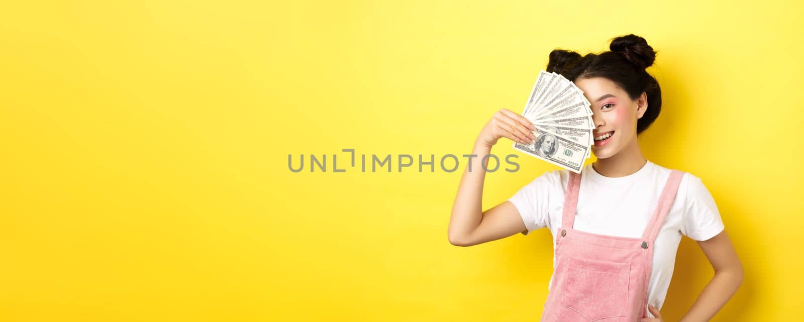 Shopping. Beautiful asian female model with glamour makeup, cover half of face with money dollar bills, smiling sensual at camera, yellow background.