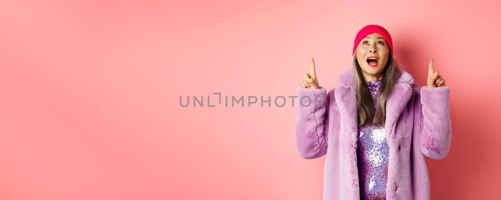 Mature asian hipster woman pointing fingers up, looking at top promo with amazement, checking out special deal, standing over pink background.