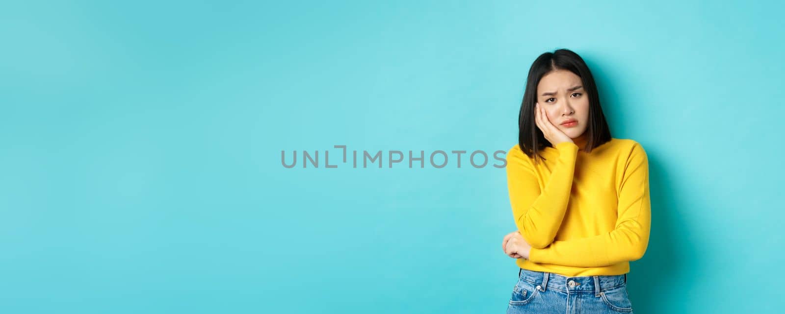 Sad and bored asian girl looking reluctant and unamused at camera, leaning face on hand, standing over blue background.