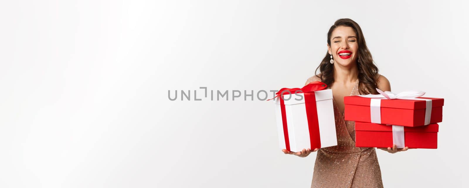 Holidays, celebration concept. Beautiful caucasian woman in elegant dress holding Christmas presents and smiling happy, standing over white background by Benzoix