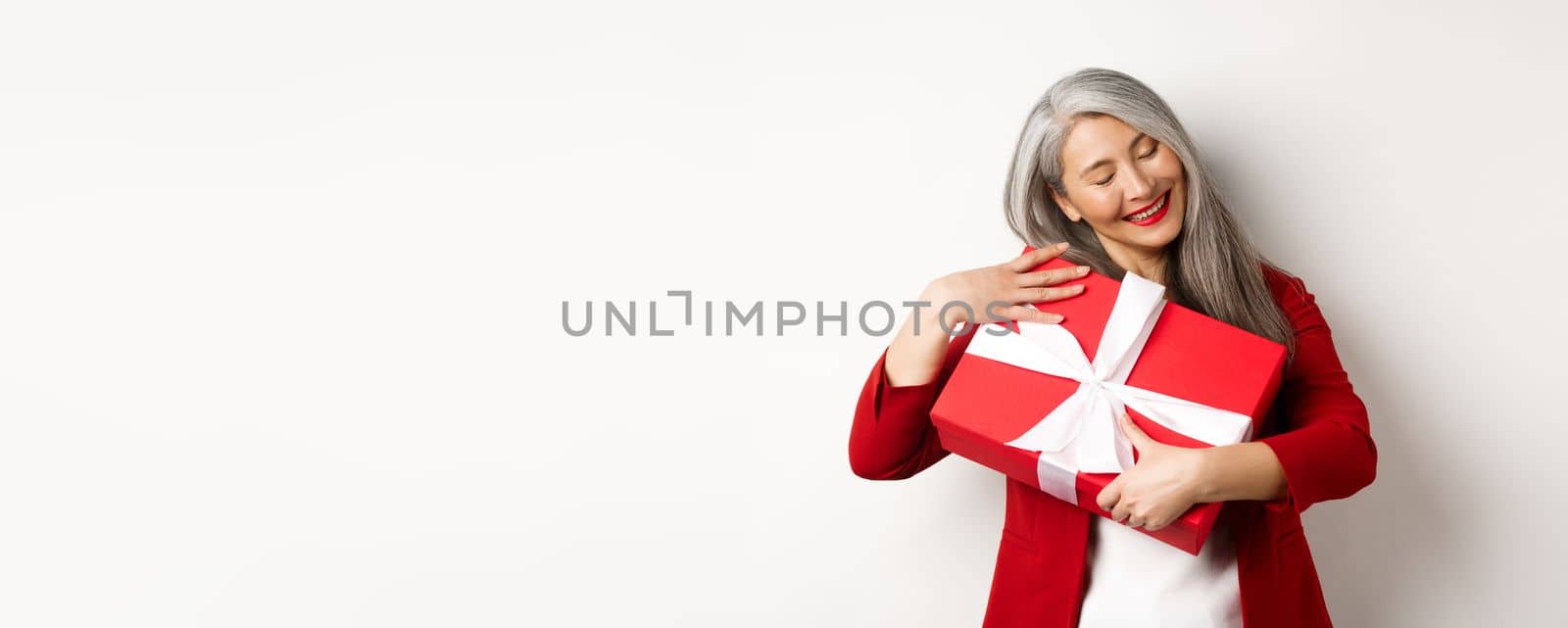 Valentine day concept. Beautiful asian senior woman hugging box with gift, smiling and looking happy, receiving present on holiday, white background.