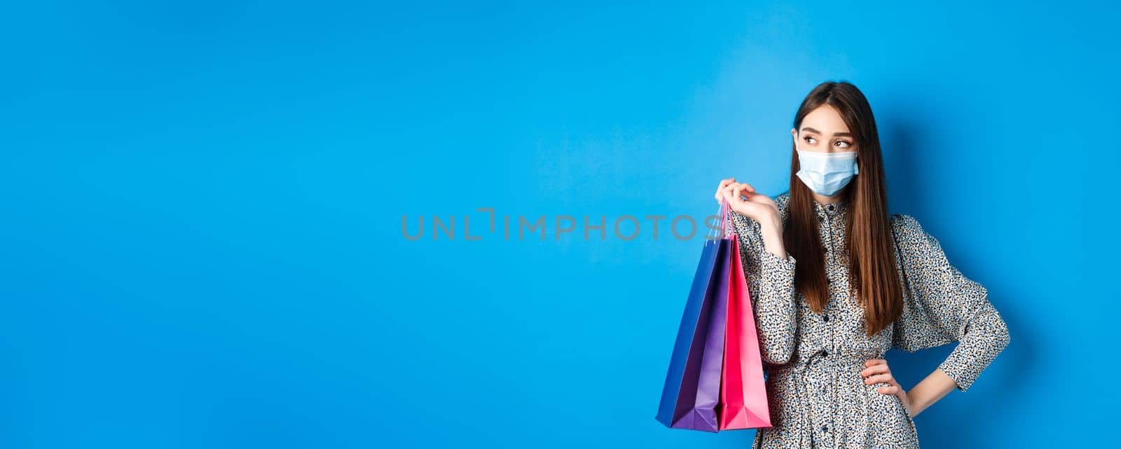 Covid-19, pandemic and lifestyle concept. Girl wearing medical mask on shopping in malls, holding paper bags and look aside at empty space, standing on blue background.