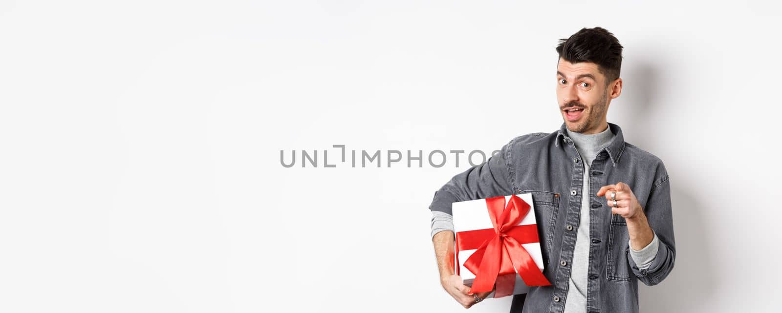 Portrait of romantic guy buying gift for Valentines day and pointing at you, inviting to special deal shopping, standing with present on white background.