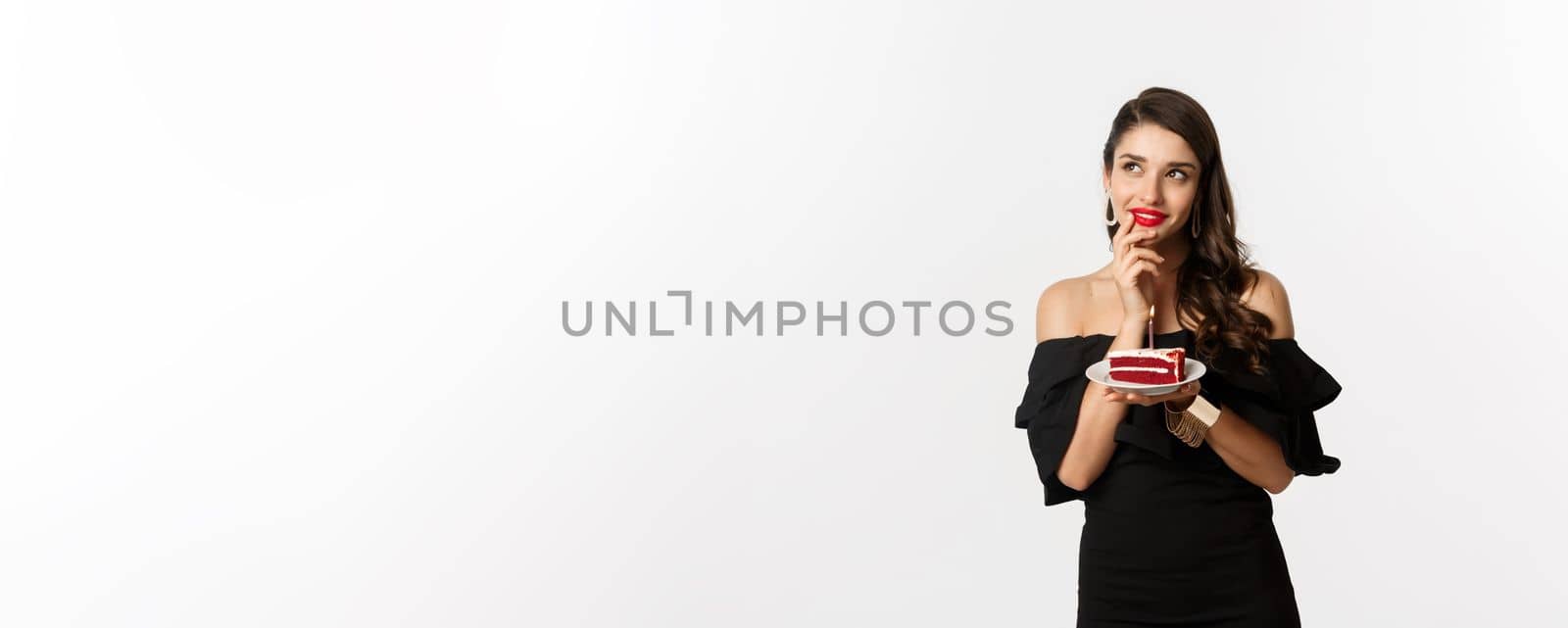 Celebration and party concept. Dreamy woman in black dress making wish, thinking and holding birthday cake with candle, standing over white background.