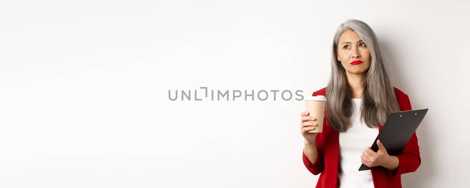 Sad asian businesswoman drinking coffee at work and looking upper left corner, standing over white background by Benzoix
