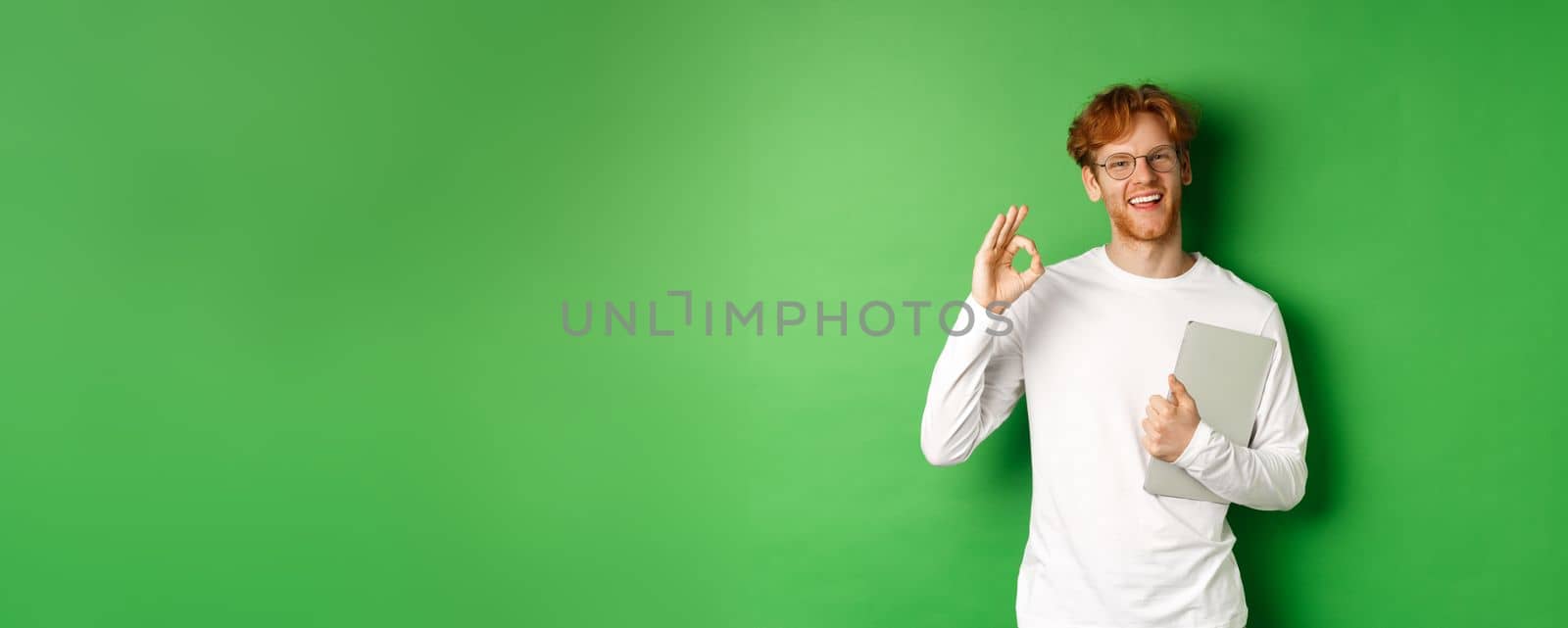 Handsome young male employee in glasses showing Ok sign, holding laptop, standing over green background.
