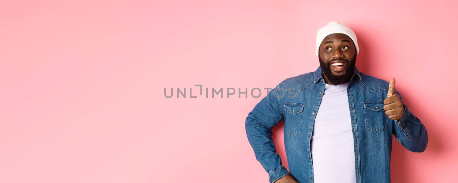 Happy Black man showing thumb-up, looking at upper left corner thoughtful, standing over pink background.