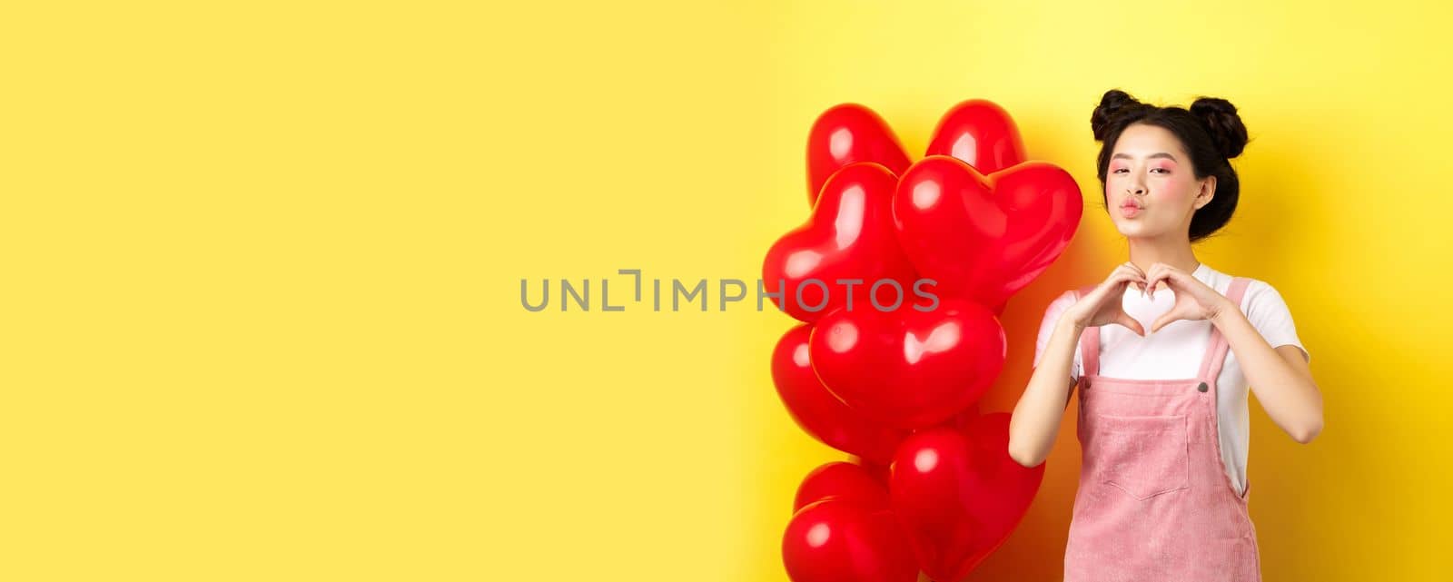 Beautiful asian woman showing heart, I love you gesture and kissing lips, standing near romantic red balloons. Concept of Valentines day.