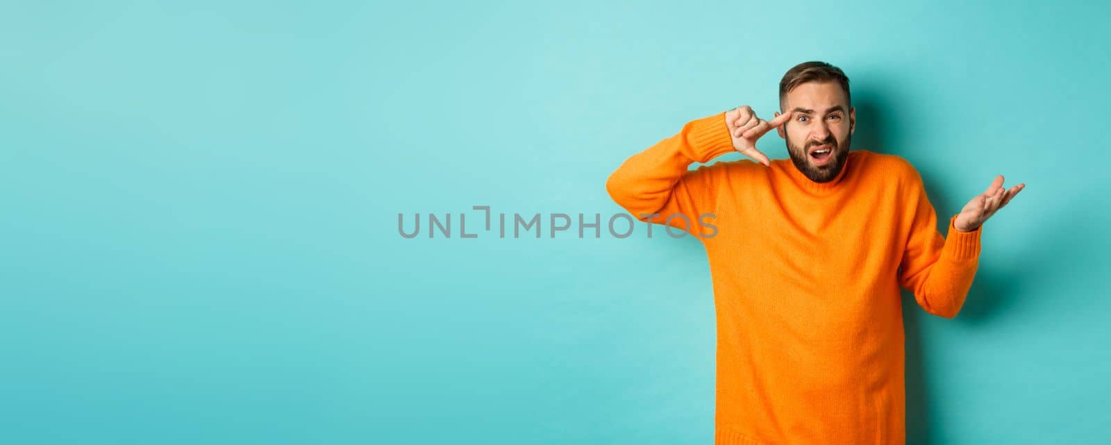 Angry man pointing at head, are you crazy gesture, scolding someone, standing over light blue background. Copy space