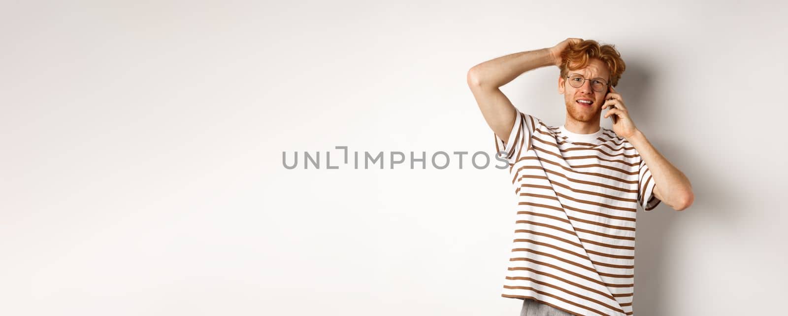 Puzzled redhead guy talking on phone, scratching head and looking confused or indecisive, standing over white background.