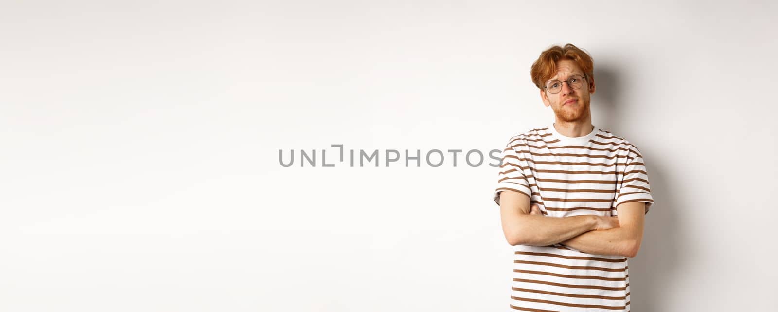 Teenage redhead guy in glasses cross arms on chest, looking skeptical and unamused at camera, standing over white background.