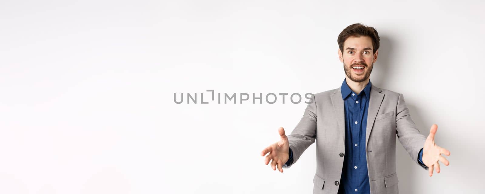 Happy handsome man in suit stretch out hands to greet business partners, welcome guests and smiling excited, standing on white background.