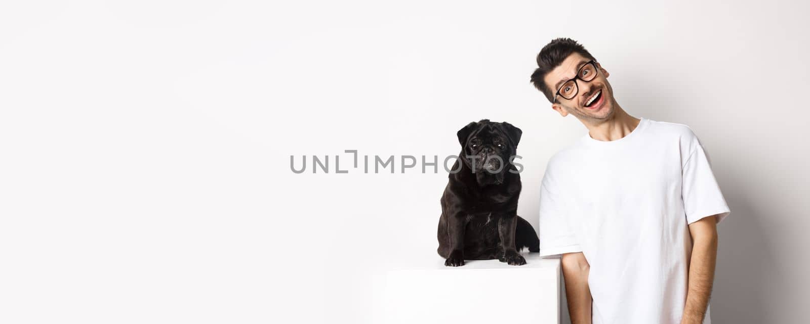 Image of handsome young man standing near cute black pug and smiling. Dog owner spending time with his pet, staring at camera happy, white background.