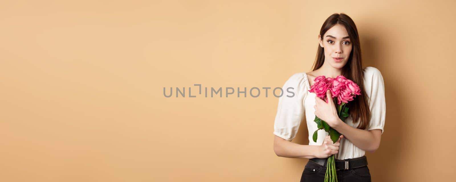 Valentines day. Surprised tender girl thanking for flowers and smiling, holding red roses and smiling grateful, standing on beige background.