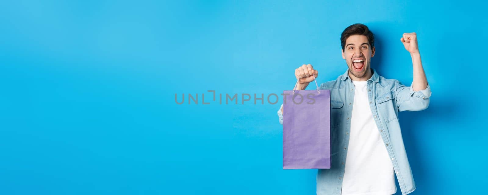Concept of shopping, holidays and lifestyle. Cheerful young man celebrating, holding paper bag and making fist pump like winner, standing over blue background.