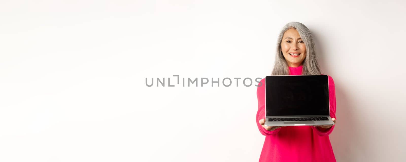 E-commerce concept. Smiling asian senior woman showing blank laptop screen and looking happy, demonstrating promo, white background.