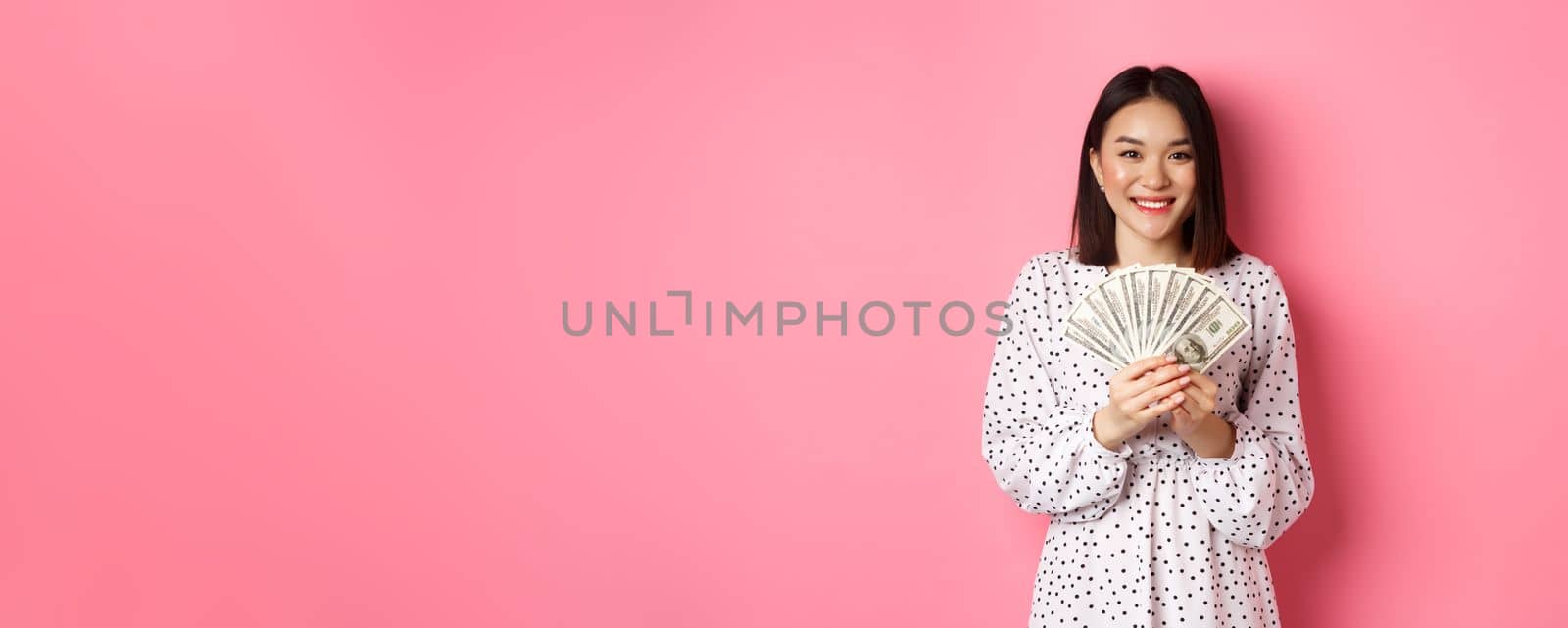 Shopping concept. Romantic asian woman in dress smiling, showing dollars money, standing over pink background.