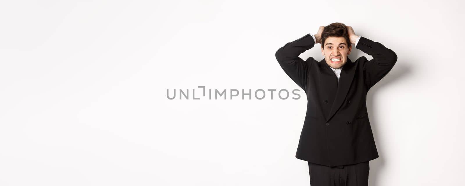 Image of frustrated and angry businessman in black suit, ripping hair on head and grimacing mad, standing tensed against white background.