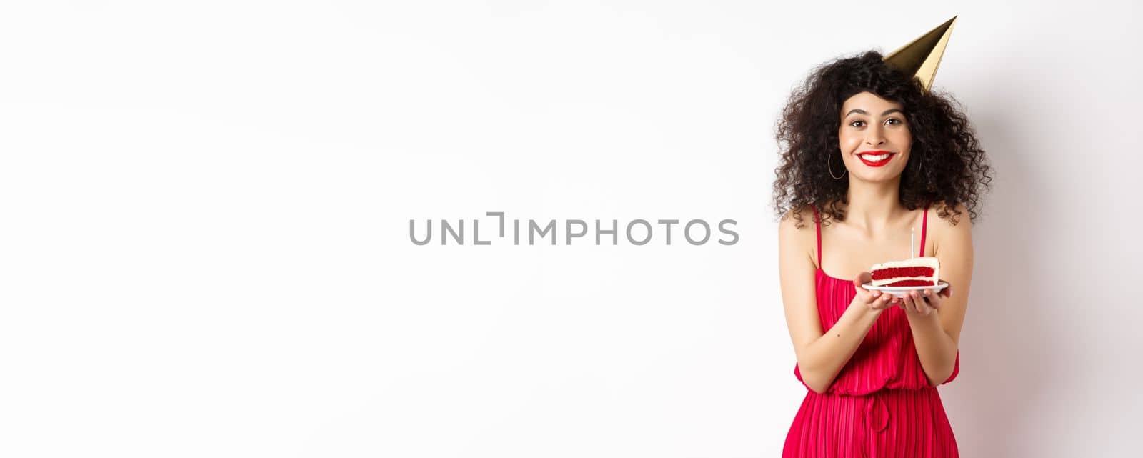 Beautiful woman in red dress, wearing party hat and celebrating birthday, holding b-day cake and making wish, smiling at camera, standing on white background by Benzoix
