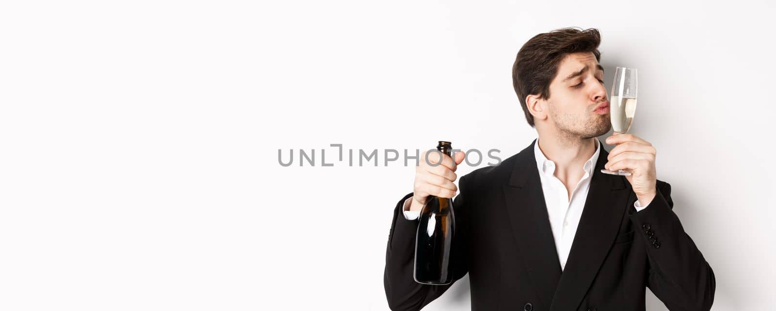 Close-up of handsome man in suit, kissing glass with champagne, getting drunk on a party, standing against white background.