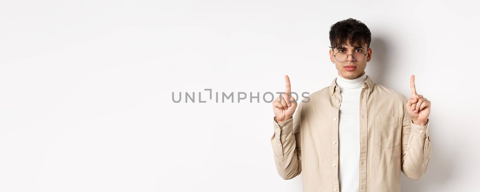 Handsome hipster guy in glasses pointing fingers up at empty space, looking serious at camera, standing on white background.