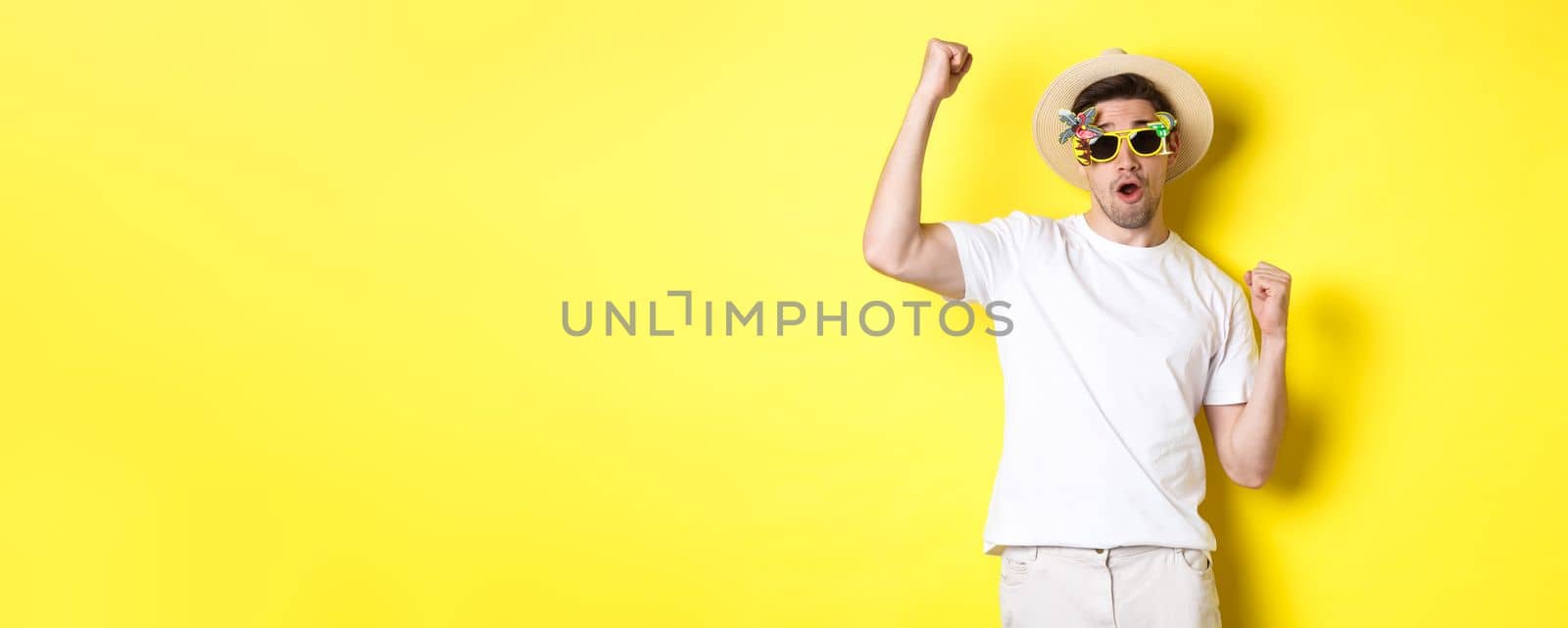 Concept of tourism and lifestyle. Happy guy tourist enjoying trip, rooting for you, fist pump and triumphing, going on journey in summer hat and sunglasses, yellow background.