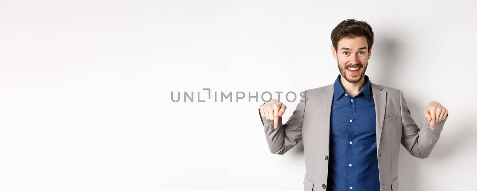 Hey check this out. Happy businessman in suit pointing fingers down and smiling, inviting to event, showing link, standing on white background.