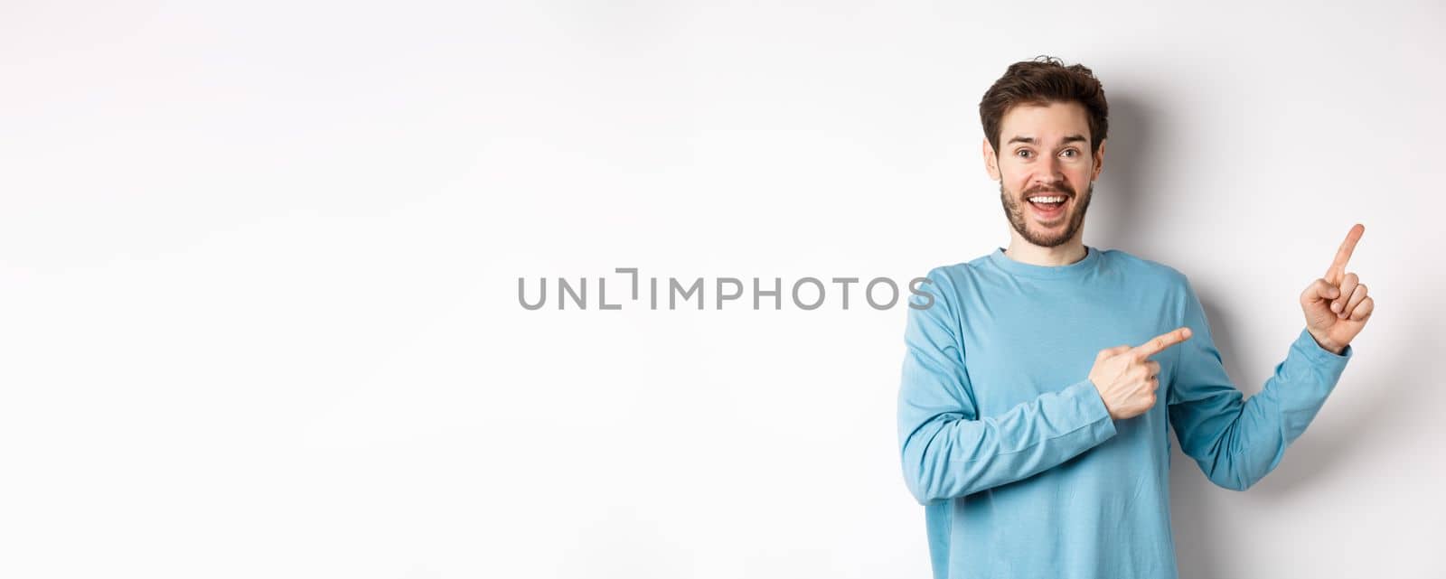 Cheerful smiling man pointing fingers right and showing banner or logo on white space for advertisements, recommending product, standing over studio background.