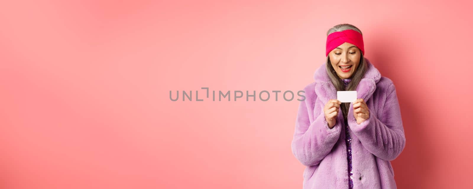 Shopping and fashion concept. Beautiful and stylish asian woman looking at plastic credit card, smiling pleased, standing over pink background.