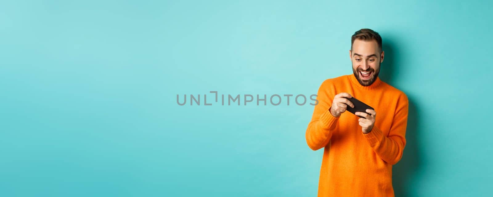 Excited adult man playing on mobile phone, looking amazed at smartphone screen, standing over turquoise background.