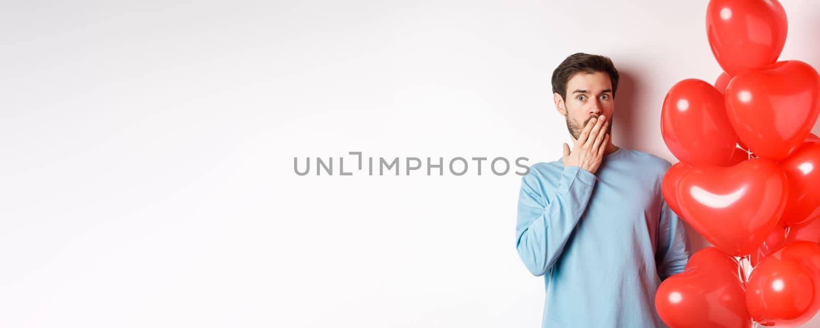 Portrait of man boyfriend standing near Valentines day heart balloons and gasping shocked, standing over white background concerned.