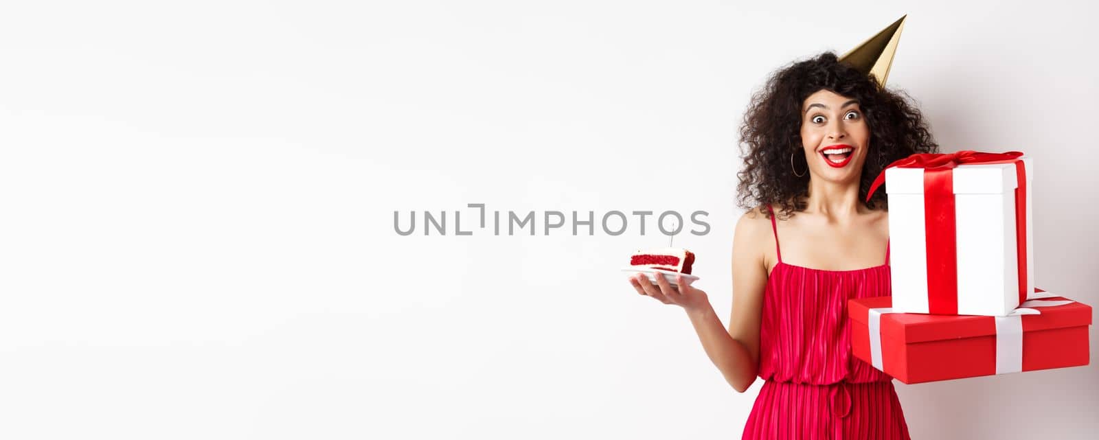 Cheerful girl in party hat and red dress, celebrating birthday, holding surprise gift and b-day cake, enjoying holiday, standing over white background.