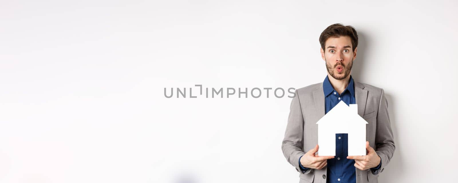 Real estate. Amazed caucasian man in suit checking out special offer for property, standing with paper house cutout and say wow at camera, white background.