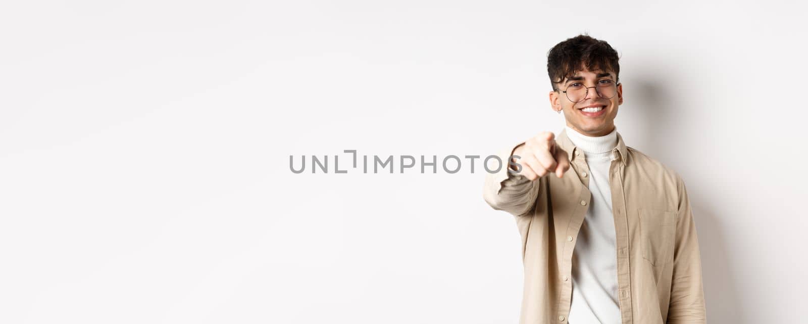 Handsome modern guy in glasses pointing at camera, smiling and choosing you, recruiting or inviting to event, standing on white background.