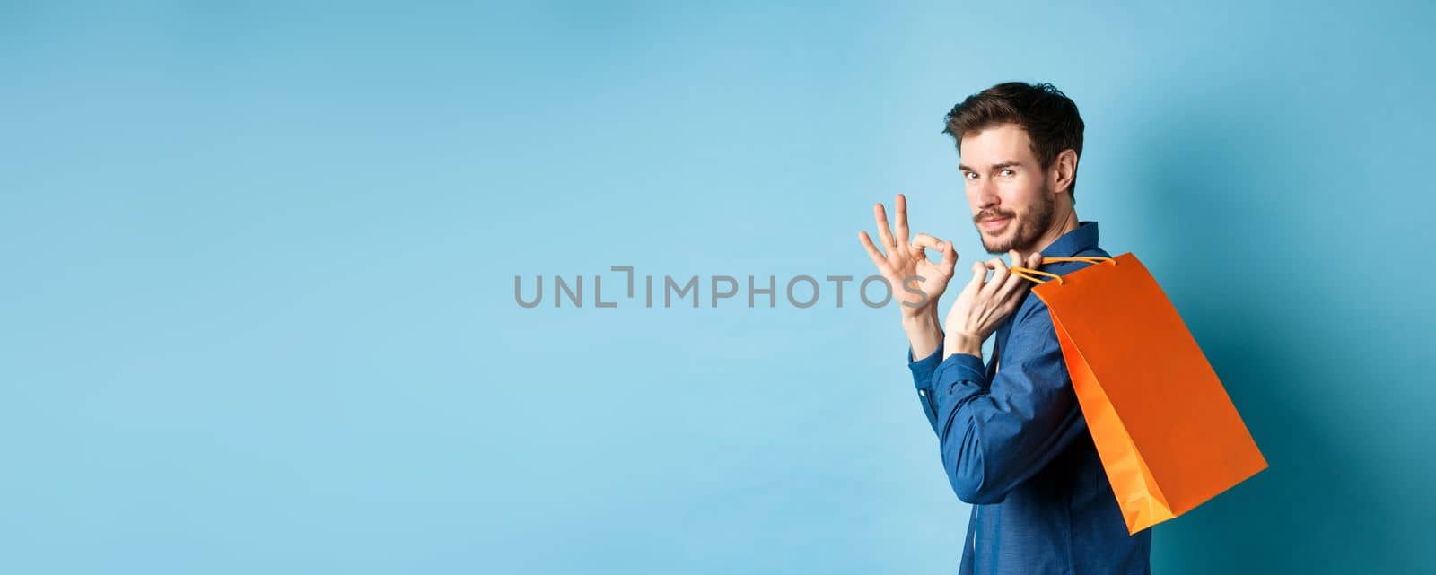 Handsome young man holding shopping bag on shoulder, turn around to look at camera and show okay gesture, recommending shop, blue background.