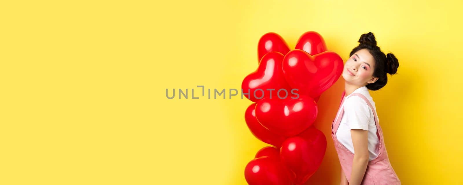 Valentines day concept. Stylish teenage asian girl posing near red hearts balloons, wear outfit for romantic date, standing happy on yellow background.