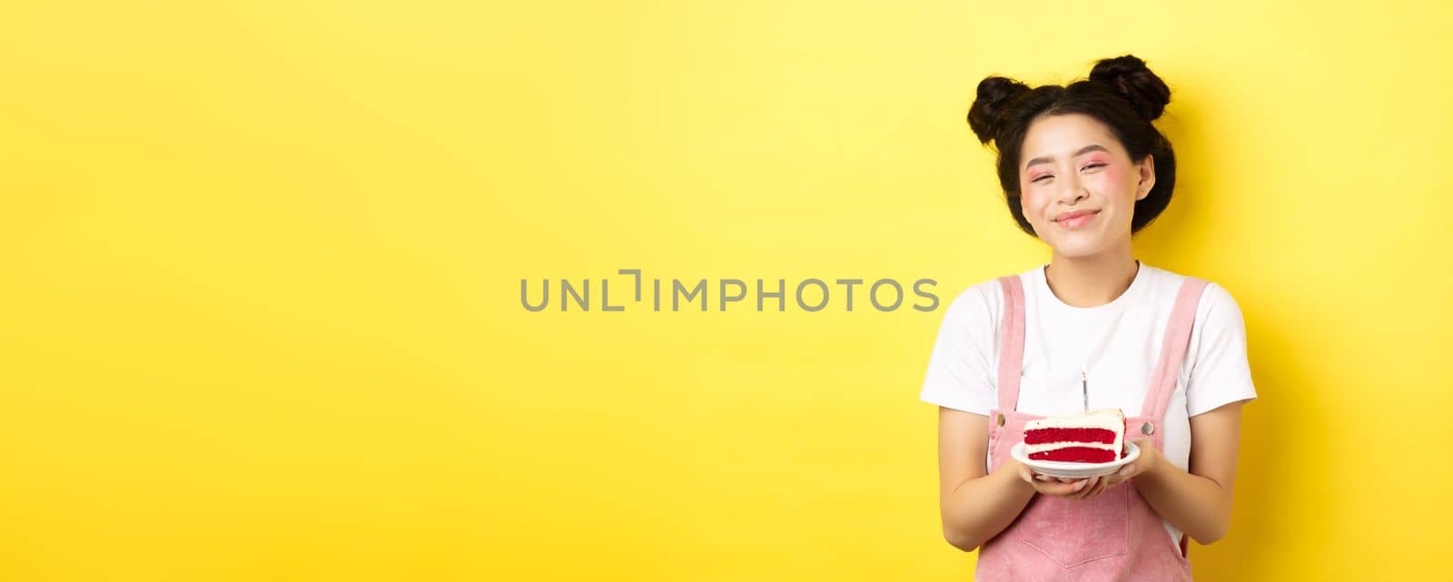 Happy asian birthday girl with bright makeup, blowing candle on cake, making wish, standing on yellow background.