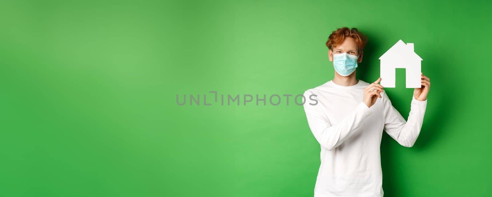 Real estate and covid-19 pandemic concept. Young redhead man in medical mask showing paper house cutout and smiling with eyes, standing over green background.