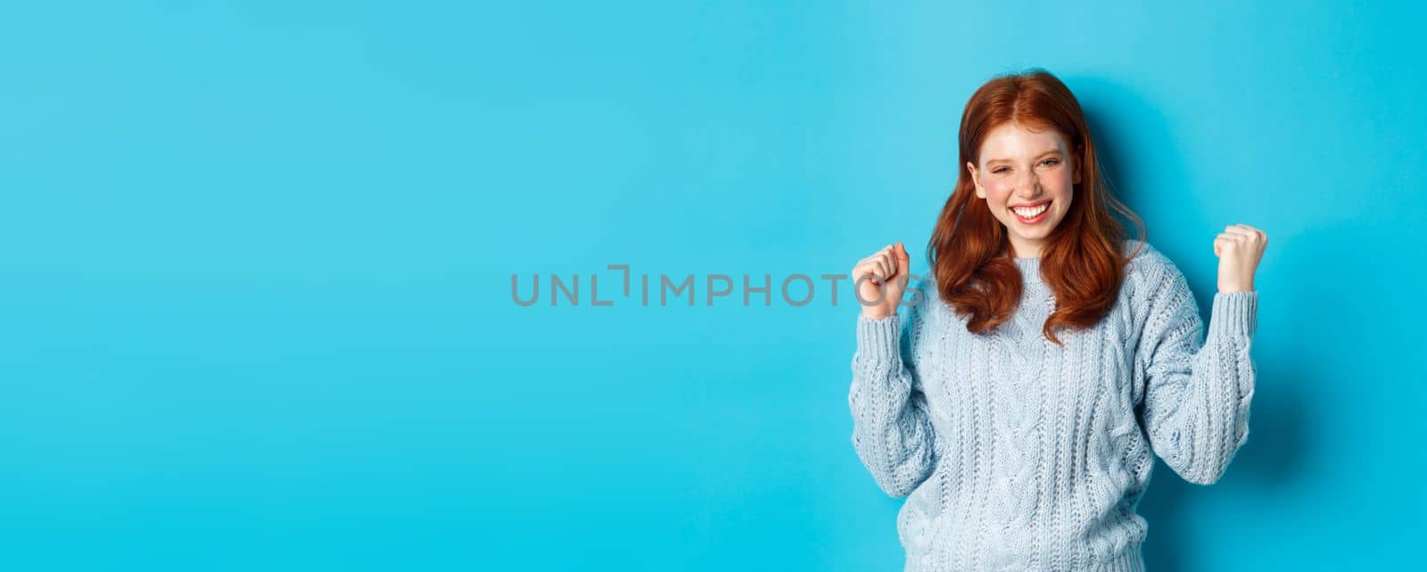 Satisfied redhead girl achieve goal and celebrating, making fist pump gesture and smiling delighted, triumphing of win, standing against blue background.