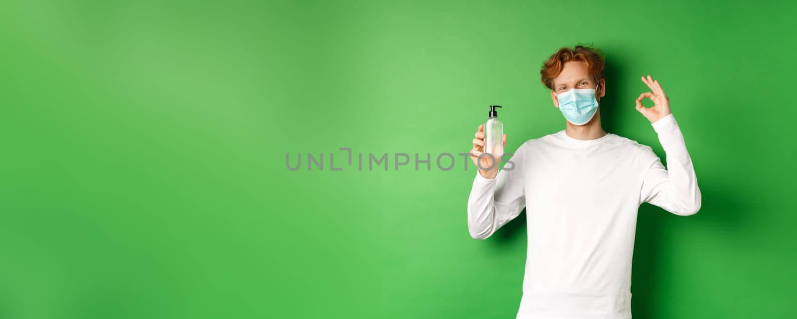 Covid-19, virus and social distancing concept. Smiling young man with red hair, wearing face mask from coronavirus, showing okay sign and hand sanitizer, green background.