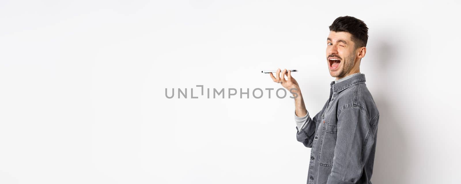 Happy guy winking at looking at camera while using voice translator app or talking on speakerphone, holding phone close to mouth, standing on white background by Benzoix