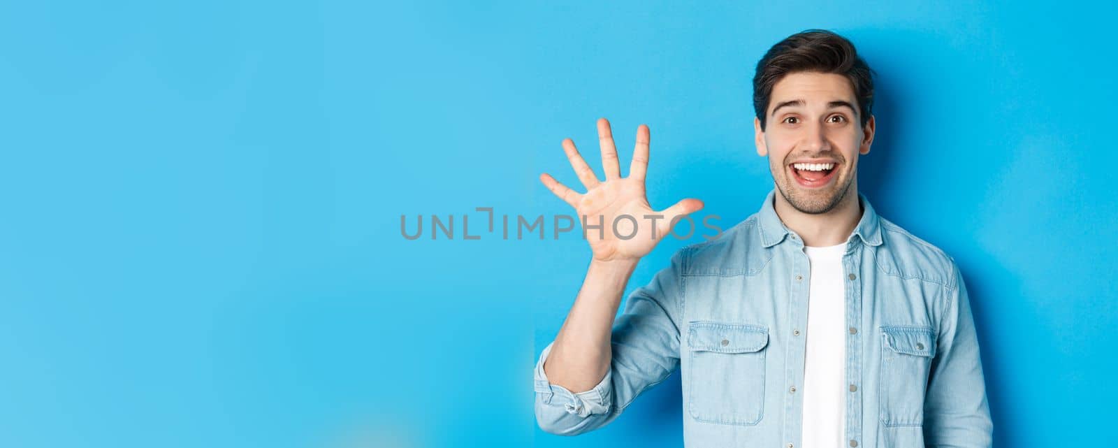 Close-up of handsome man smiling, showing fingers number five, standing over blue background.