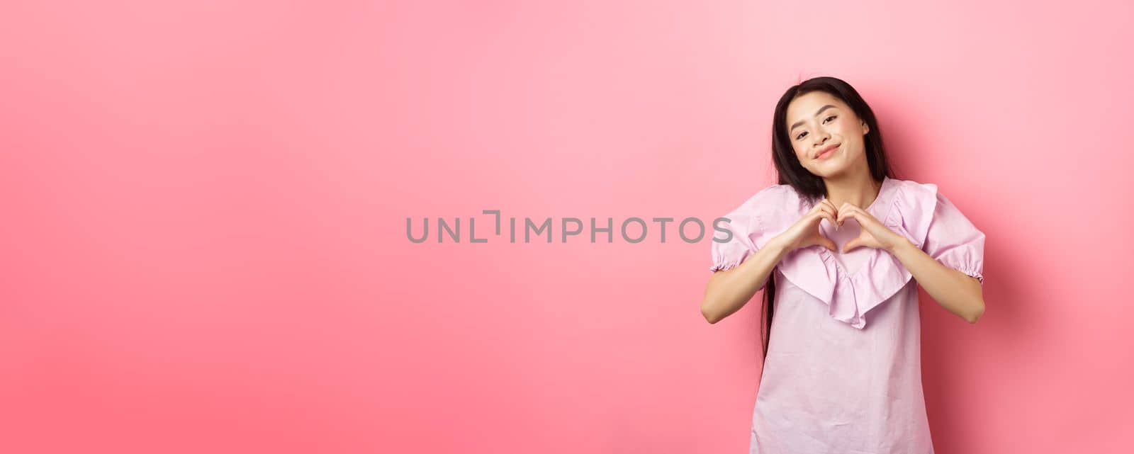 Valentines day concept. Beautiful korean teenager in dress showing heart gesture and smiling with admiration and affection, falling in love, feel romantic, pink background by Benzoix