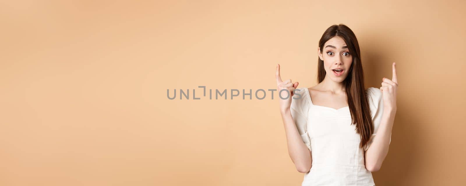 Wow look here. Amazed cute girl making announcement, pointing fingers up and look excited, showing promo offer, beige background.