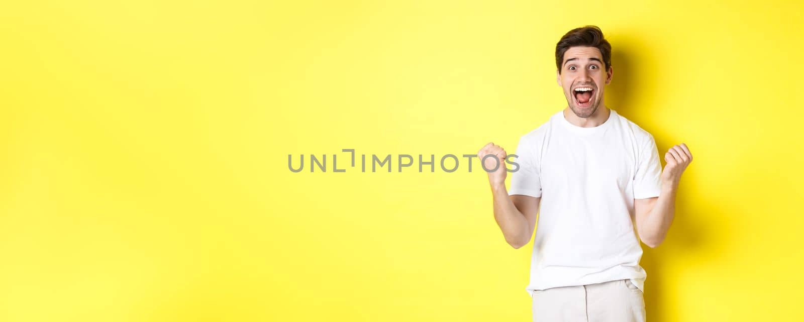 Excited and lucky man winning, clench fists and looking happy, triumphing and celebrating, standing over yellow background.