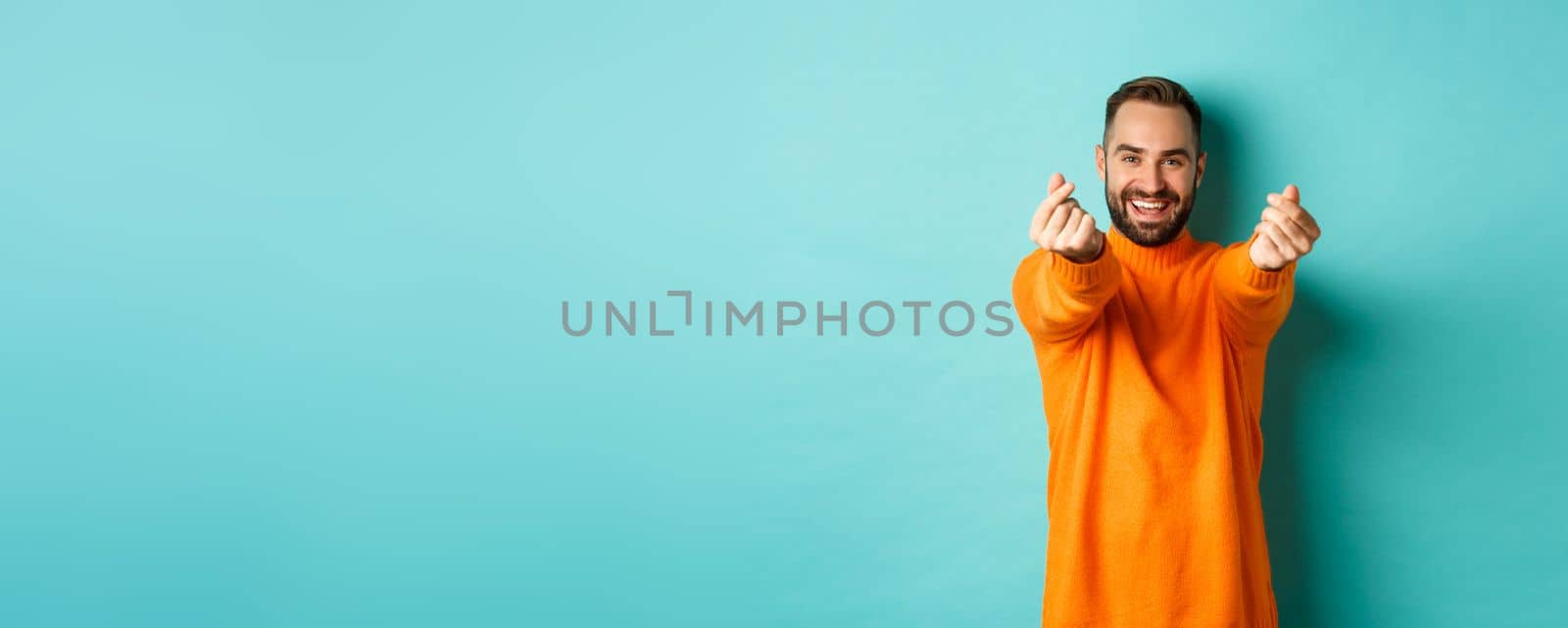 Smiling handsome man showing hearts and looking happy, lucky gesture, standing in orange sweater over light blue background by Benzoix