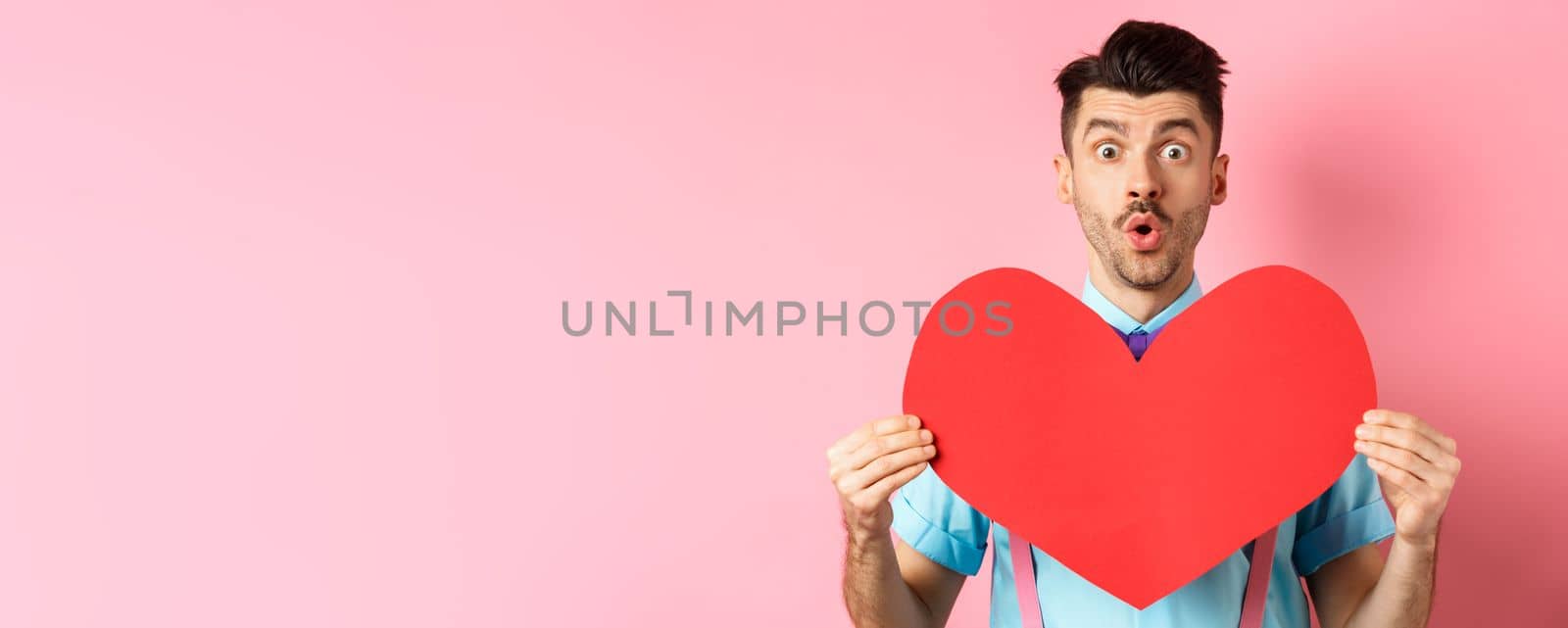 Valentines day concept. Amused young man with bow-tie and moustache, showing heart cutout and looking for love, standing on pink romantic background.