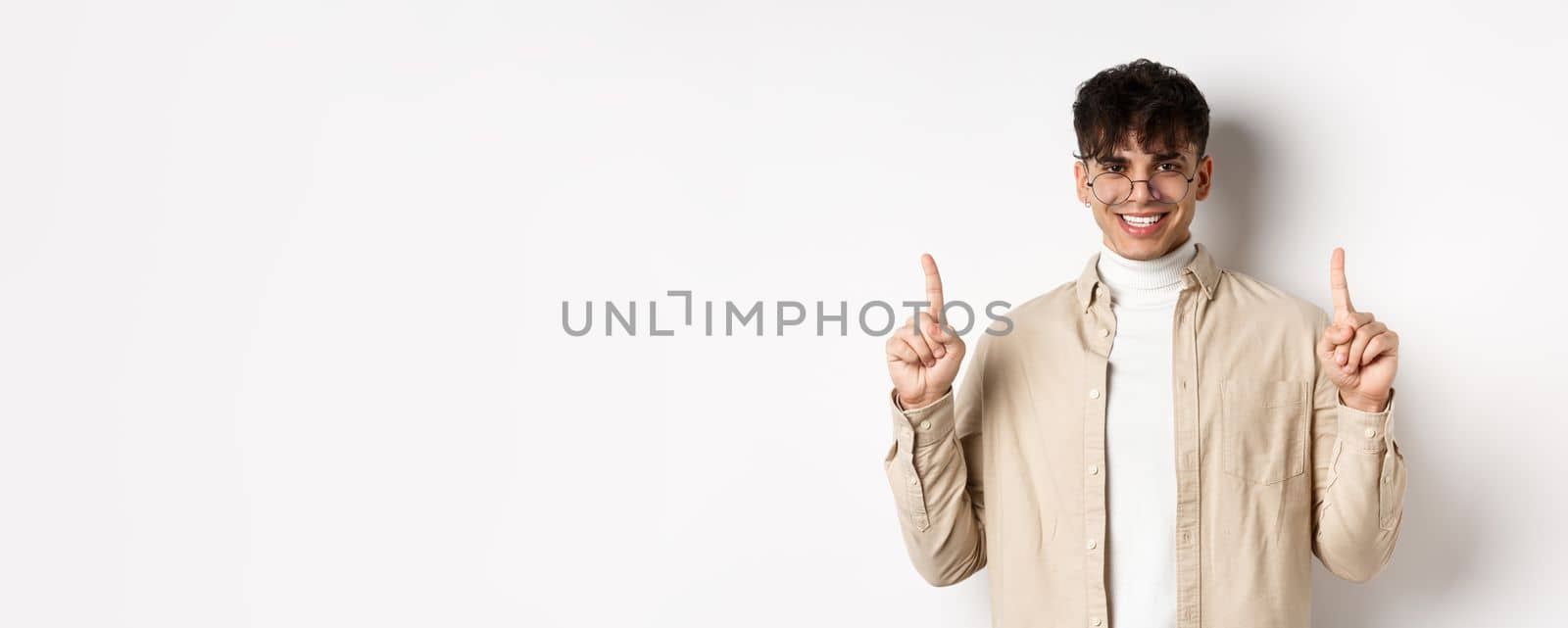 Portrait of handsome hipster guy in glasses showing advertisement and smiling, pointing fingers up at top offer, standing on white background by Benzoix