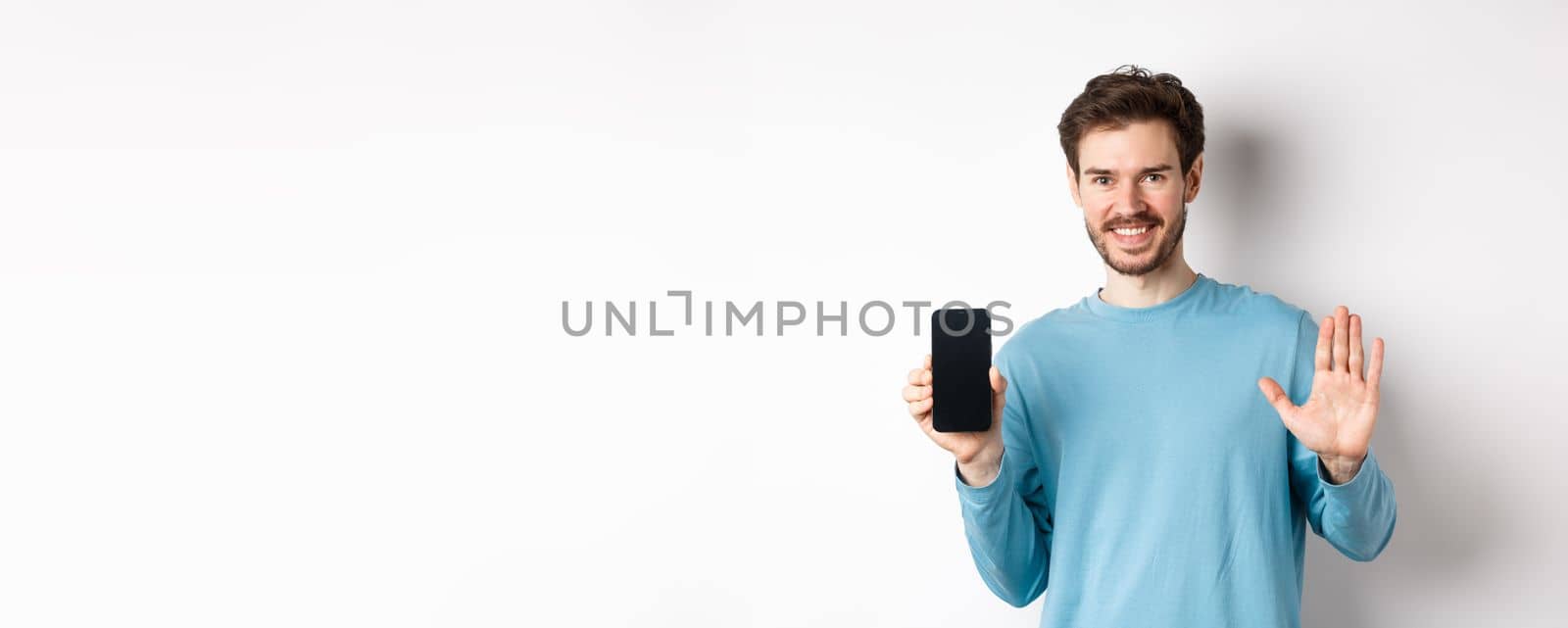 Handsome caucasian man with beard, showing empty smartphone screen and number five, raising hand to wave and say hello, standing on white background by Benzoix